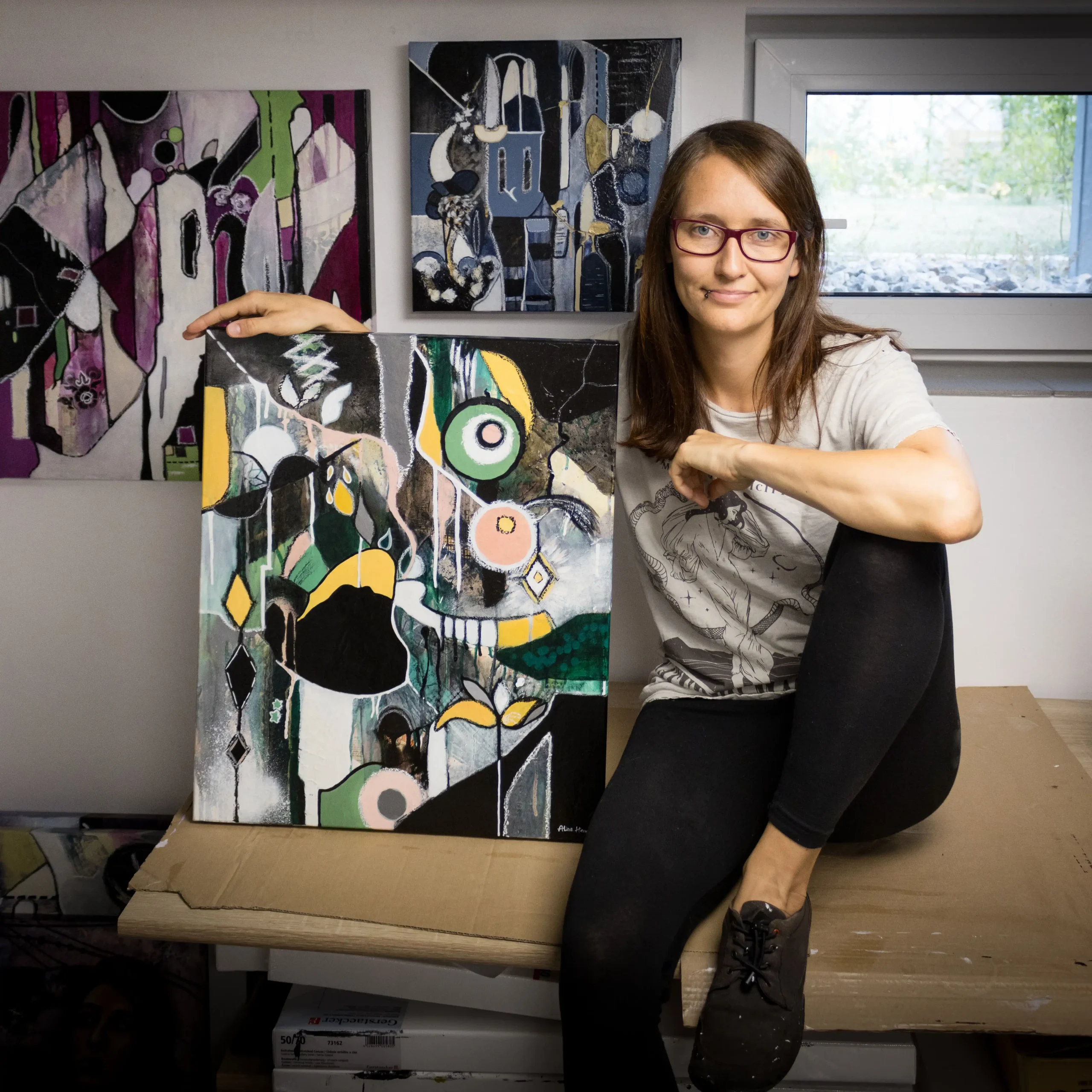 Abstract mixed media and oil artist Alina Hermann sitting on a table with three of her artworks.